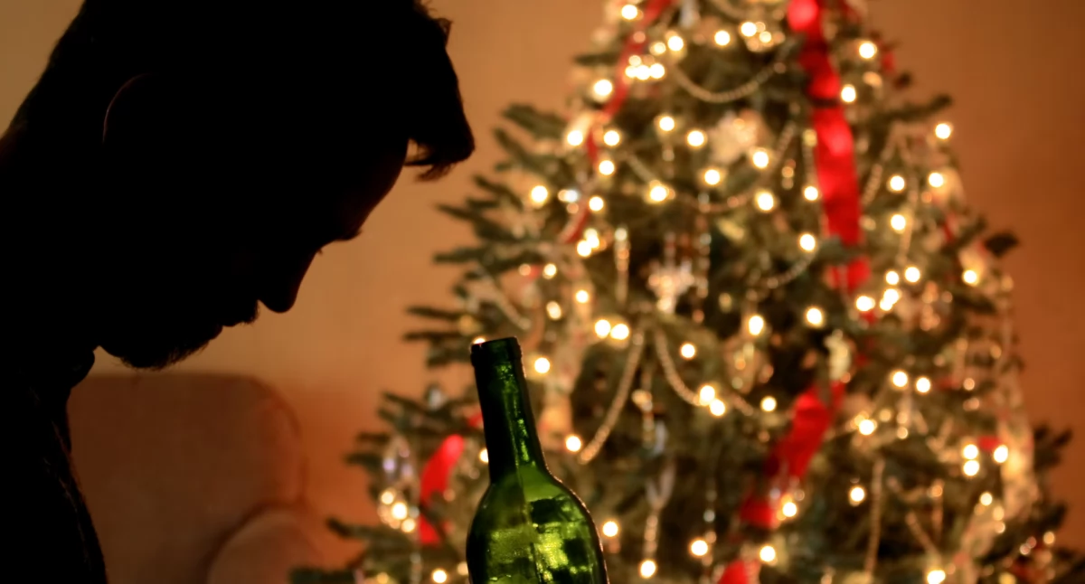 Mężczyzna ze spuszczoną głową pijący alkohol na tle choinki ze świątecznymi ozdobami/ canva