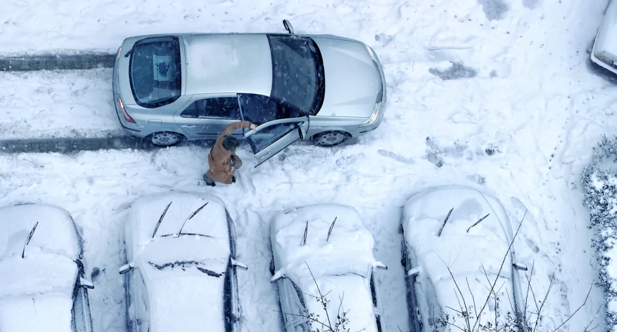 Zaśnieżone samochody na parkingu i wsiadający do jedno z nich pasażer/podróżny. Fot.: Canva