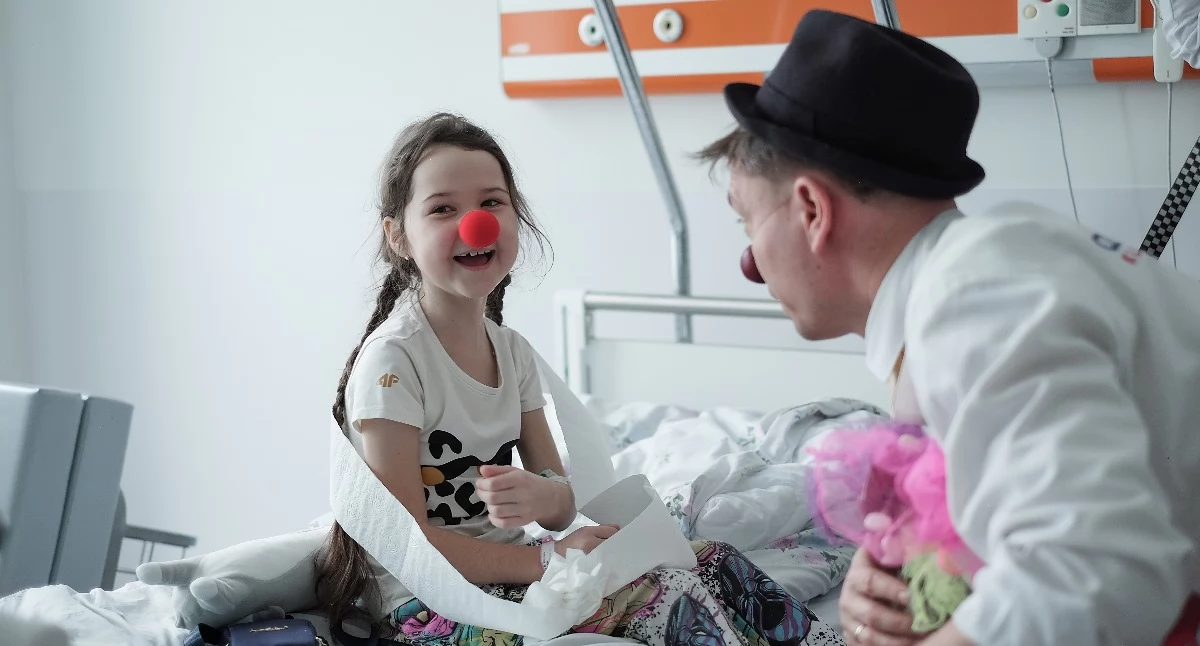 Na zdjęciu: uśmiechnięta dziewczynka w szpitalnym łóżku i klown medyczny /fot. Karolina Matuszewska.